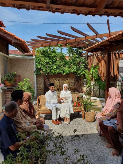 Tetamu berkumpul di bangku rotan di bawah pergola hijau.